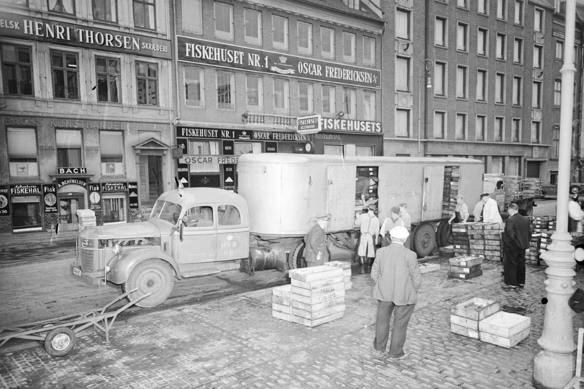 København 1951: Der læsses frisk fisk af i Indre By. En tidlig morgen - der gøres klar til dagens handel. Skagensbanen og Danske Statsbaners fragtrute for fisk Skagen-Grenå-København-Hundested udlæsser gods ved fiskemarkedet på Gammel Strand i København. I baggrunden ses nogle af datidens fiskerestauranter i gaden. Billedet er taget før solopgang kl. 05.10, og der er derfor benyttet blitz ved optagelsen. (Foto: Danmarks Jernbanemuseum)